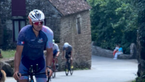 A cyclist in a blue and pink jersey rides uphill on a road with stone buildings and greenery in the background, followed by three other cyclists.
