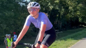 A cyclist wearing a white helmet, sunglasses, a light purple jersey, and black shorts rides a bike on a sunny day, with trees and people standing on the grass in the background.