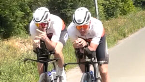 Two cyclists in matching white helmets and jerseys ride closely side by side on a paved road, leaning forward in an aerodynamic position on their bikes. Green foliage lines the road in the background.
