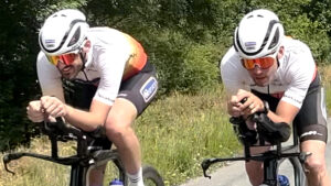 Two cyclists in matching white jerseys and helmets ride side by side on a road, leaning forward on their handlebars in an aerodynamic position, with greenery in the background.
