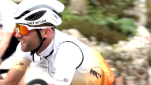 A cyclist wearing a white helmet, sunglasses, and a white and orange Café Vitesse jersey rides outdoors, smiling during a race or event. The background is blurred greenery.