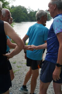 A group of older adults in athletic clothes stand on a gravel path by a lake, chatting and smiling, with two of them gently holding hands in the center of the image. Trees and water are visible in the background.