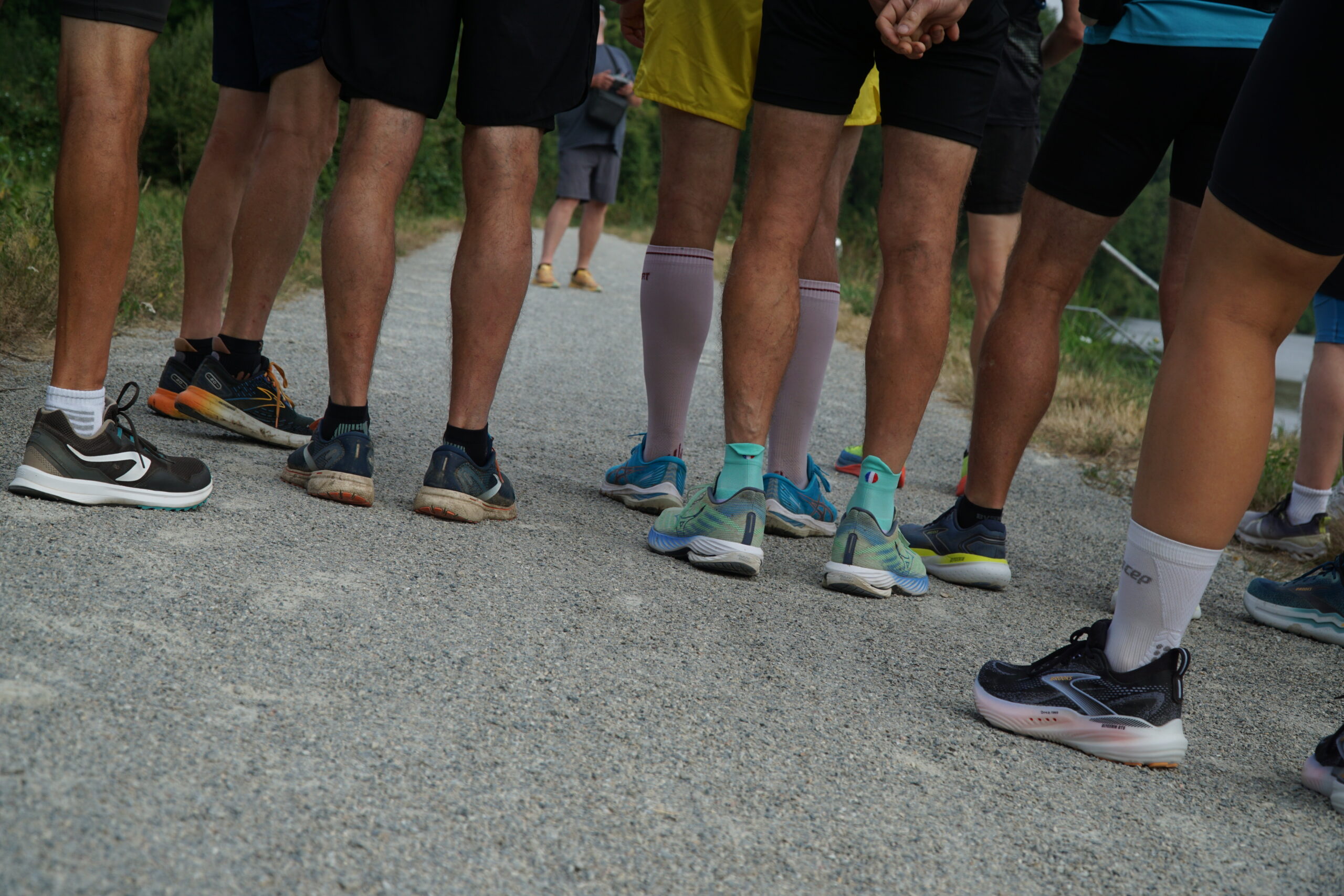A group of people standing on a gravel path, shown from the knees down, wearing athletic shoes and shorts, appearing to prepare for a run or outdoor activity.