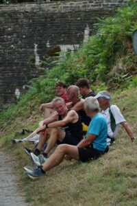 Six men in athletic clothing sit on a grassy slope, smiling and laughing together by a stone wall and green foliage, enjoying a relaxed moment after exercise at a glorious chateau in the Loire. The atmosphere is casual and full of camaraderie.