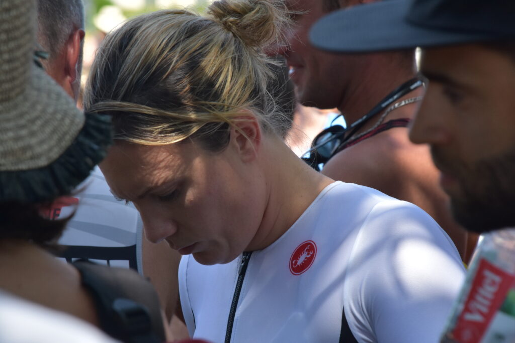 A woman in a white athletic outfit with a red logo looks down, surrounded by people outdoors near a glorious chateau in the Loire. The image is candid and focused on her, with blurred individuals in the foreground and background.