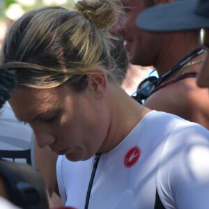 A woman in a white athletic outfit with a red logo looks down, surrounded by people outdoors near a glorious chateau in the Loire. The image is candid and focused on her, with blurred individuals in the foreground and background.