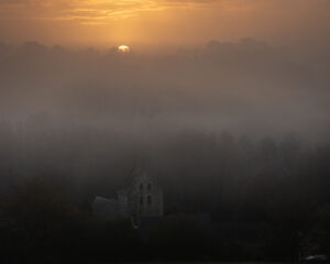 A glorious chateau in the Loire sits among trees shrouded in morning fog, with the sun rising above the misty horizon, casting an orange glow across the sky.