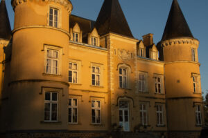 A large yellow chateau with tall, conical towers, multiple white-framed windows, and ornate architectural details, bathed in warm sunlight.