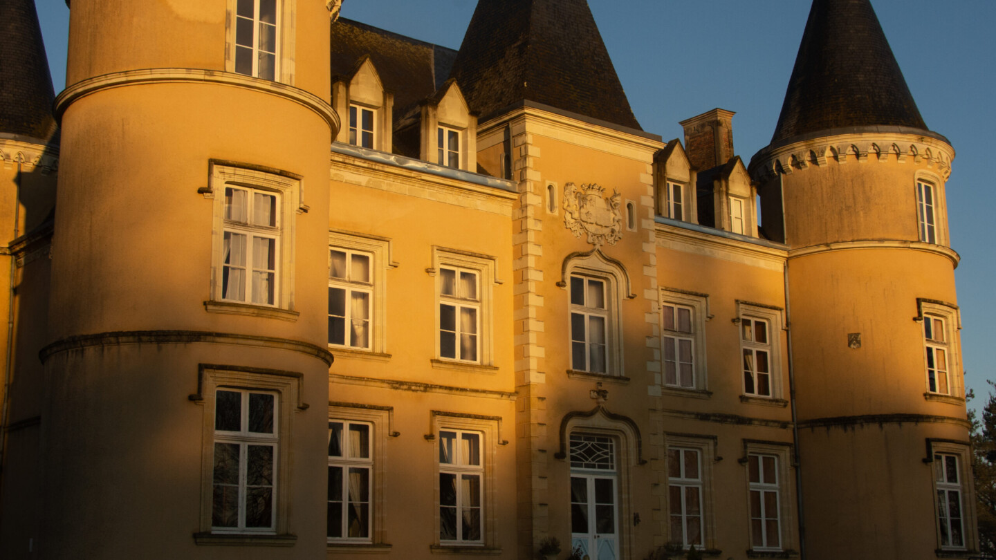 A large yellow chateau with tall, conical towers, multiple white-framed windows, and ornate architectural details, bathed in warm sunlight.