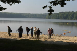 A group of people with backpacks and walking sticks stand on a sandy lakeshore near calm water, recalling adventures by the Glorious Chateau in the Loire, with trees in the background and yellow buoys floating in the lake.