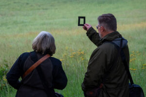 Two people with shoulder bags stand in a grassy field near a glorious chateau in the Loire. One person holds up a small rectangular black frame, looking through it towards the landscape. Both are facing away from the camera.