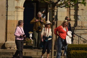 Five women with cameras stand and walk outside a glorious chateau in the Loire on a sunny day. Some wear hats and sunglasses, while one woman in the foreground has a coral sweater and white jacket. Greenery and a tree are nearby.
