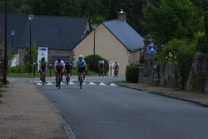 A group of cyclists ride up a quiet, tree-lined street in a small village with stone houses and greenery on either side. The sky is overcast and the road has a speed bump ahead.