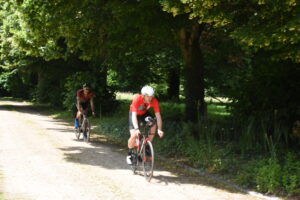 Two cyclists ride on a sunlit dirt path through a green forested area. The leading cyclist wears a red jersey and white helmet, while the second cyclist follows behind in a patterned jersey, both focused on the ride.