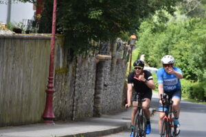 Two cyclists ride on a narrow road next to a stone wall and greenery. Both are wearing helmets and sunglasses; one in black, one in blue. They are giving thumbs up and appear to be enjoying their ride.