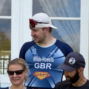Three athletes pose in front of glass doors. The central man wears a white cap, sunglasses on his head, and a blue racing suit labeled GBR. A woman with sunglasses and a bearded man in a cap stand beside him.