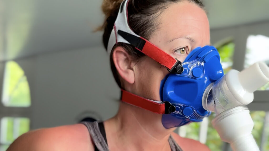 A person wearing a blue and red respiratory mask with white straps is indoors, connected to a tube, possibly performing a breathing or medical test. The background is softly lit with windows.