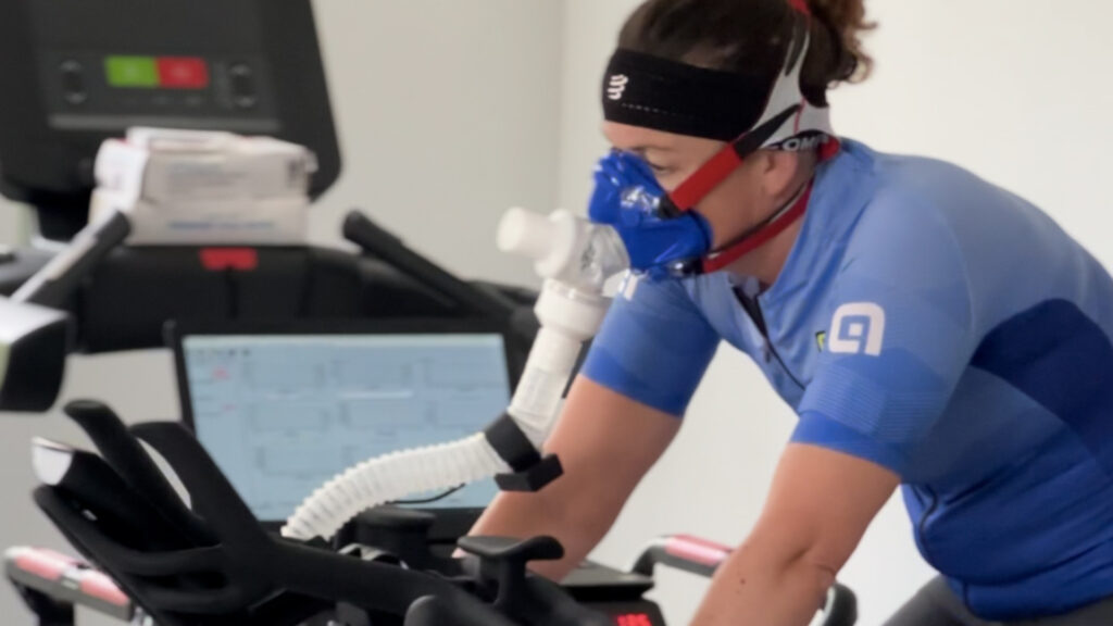 A person wearing a blue cycling outfit and a respiratory mask rides a stationary bike indoors, with a treadmill and a computer monitor displaying data in the background.