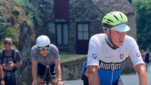 Two cyclists, one wearing a green helmet and Bigfoot jersey, ride on a sunny day, followed by another cyclist in a white helmet. In the background, two people stand near a stone building.