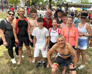 A group of smiling people, some in triathlon gear and some in casual clothes, pose together outdoors on grass at a busy event with a crowd and finish line in the background.