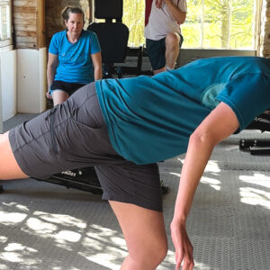 A woman in athletic clothes balances on one leg with her arms extended forward at a gym. Two other people are in the background, one sitting and one standing, near exercise equipment and large windows.