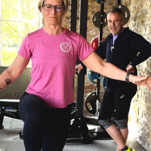 A woman in a pink shirt balances on one leg in a gym, while two men in athletic clothing stand behind her near weights and exercise equipment, smiling. Sunlight streams in through a window.