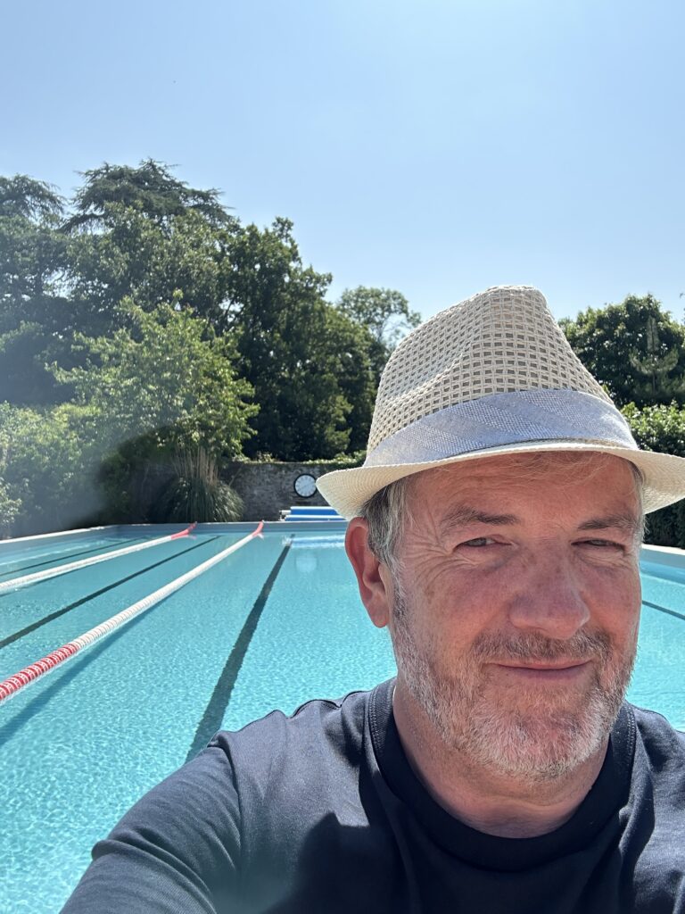 A man wearing a light-colored hat and navy shirt smiles at the camera in front of an outdoor swimming pool with lane dividers, surrounded by trees and greenery under a clear blue sky.