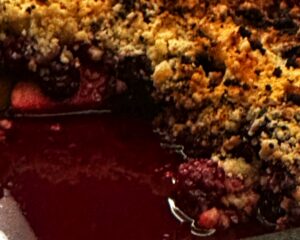 A close-up of a fruit crumble dessert with a golden-brown, crumbly topping and vibrant red fruit filling visible underneath.