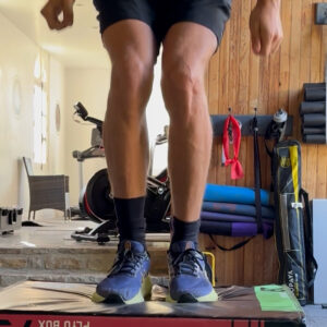A person in athletic wear and blue sneakers is mid-jump onto a plyo box in a home gym with exercise equipment and yoga mats in the background.