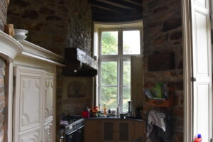 A rustic kitchen with stone walls, a large window letting in daylight, wooden shelves, a black oven, various bottles and kitchen items on the counter, and cream-colored cabinets.