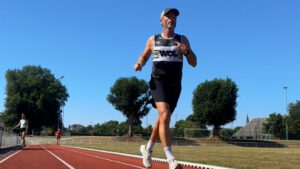 A man in athletic gear and a cap jogs on a red running track under a clear blue sky, with green trees and a grassy field in the background. Two other people are visible further along the track.
