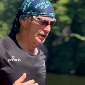 A man wearing sunglasses, a headscarf, and a black parkrun shirt runs outdoors on a sunny day, with trees and greenery blurred in the background.