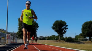 A runner in a bright yellow tank top and sunglasses jogs on a red track under a clear blue sky, with three other runners approaching in the background near trees and grassy areas.