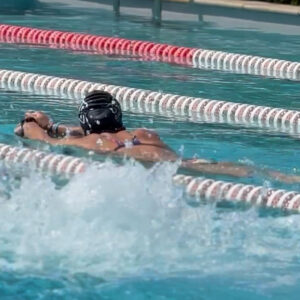 A swimmer in a pool wearing a swim cap and goggles performs the breaststroke in a lane, while water splashes nearby, possibly from another swimmer. The pool is divided by red and white lane dividers.