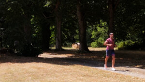 A man in a pink shirt and blue shorts jogs on a sunlit path through a park with tall trees, casting shade over part of the walkway. Dry grass covers the ground.