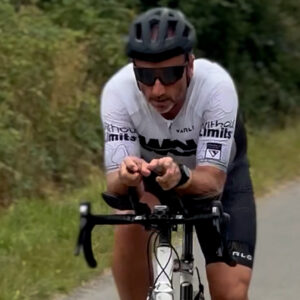 A cyclist wearing a helmet, sunglasses, and a white jersey rides on a road bike in an aerodynamic position, with trees and greenery in the background.