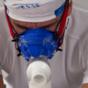 A person wearing a white shirt, headband, and a blue respiratory face mask is focused downward, possibly engaged in a physical or medical activity.
