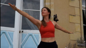 A woman wearing a red sleeveless top and black pants practices a yoga pose with arms extended outside a glorious chateau in the Loire, framed by large windows and cream-colored walls.