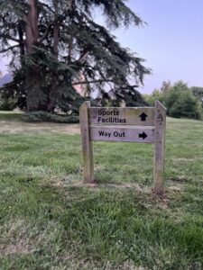 A wooden sign on grass points left for Sports Facilities and right for Way Out, set amid large trees and greenery—evoking the tranquil grounds of a glorious chateau in the Loire.