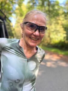 An older woman with light hair tied back smiles outdoors. She wears sporty sunglasses and a zipped-up athletic top. The background is a sunny, tree-lined path in a park or forest.