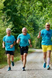 Three older men jog on a tree-lined gravel path. All wear blue shirts, and one wears yellow shorts. They smile and make peace signs, appearing cheerful and energetic during their run outdoors.