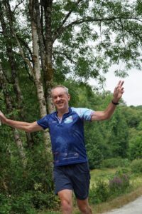 A man wearing a blue athletic shirt and shorts runs on a path in a green, wooded area, smiling and holding his arms out wide as if celebrating or balancing. Trees and foliage surround him.