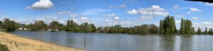 Panoramic view of a calm lake with sandy shore on the left, surrounded by green trees under a blue sky with scattered clouds.