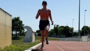 A person wearing a red sports bra, black shorts, and a cap runs on a sunny outdoor track, with trees and structures in the background.