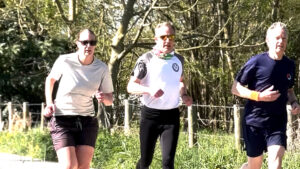 Three adults are jogging outdoors on a sunny day, surrounded by greenery and trees. Two men and one woman appear focused as they run along a path, wearing sports clothing and sunglasses.