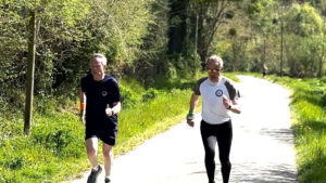 Two men are running on a sunlit paved path surrounded by trees and greenery. One wears a navy outfit, the other wears a white shirt and black leggings. Both appear focused as they run outdoors.
