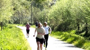 Four people are jogging on a sunlit path surrounded by green trees and grass. The runner in front wears sunglasses and a light shirt; the others follow behind at varying distances.