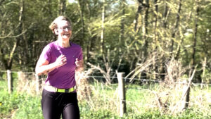 A smiling person with short hair runs outdoors on a sunny day, wearing glasses, a purple shirt, and black leggings. Green trees and a wire fence are visible in the background.