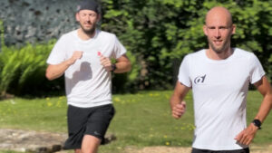 Two men in white shirts and black shorts are jogging outdoors on a sunny day, with greenery and a stone path in the background. The man in front is smiling while the man behind wears a cap and has a serious expression.