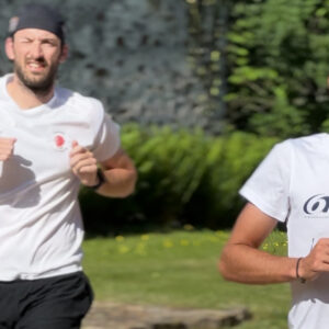 Two men in white t-shirts are jogging outdoors on a sunny day. One is in the foreground smiling, while the other is slightly behind and appears focused. Greenery and a stone wall are visible in the background.